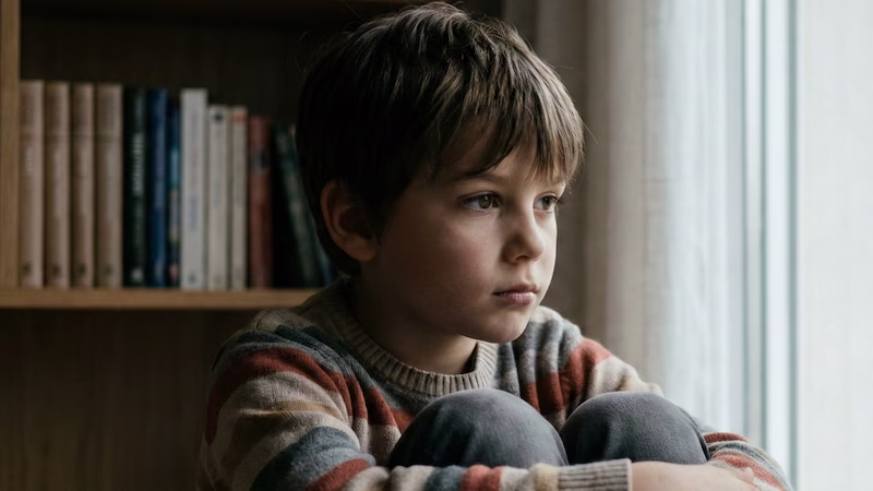 Niño mirando por la ventana con expresión triste.