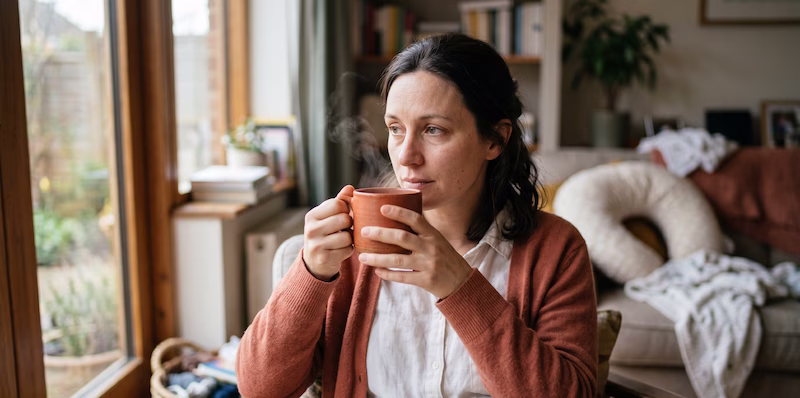 Mujer sosteniendo una taza de té, mirando por la ventana con expresión pensativa, reflejando la necesidad de autocuidado psicológico en el postparto.