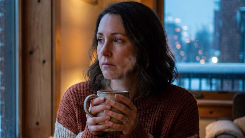 Persona mirando por la ventana con expresión pensativa, contrastando con la decoración navideña difuminada.