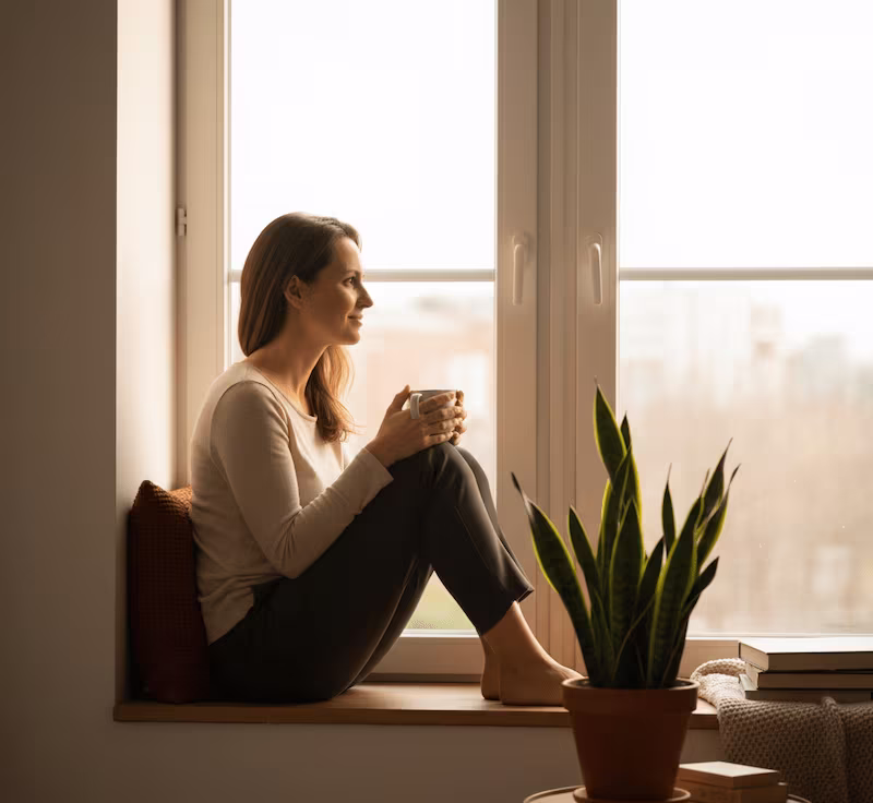 Mujer sentada en una ventana, mirando hacia afuera con una expresión de calma y autoconfianza, simbolizando el trabajo de la autoestima.